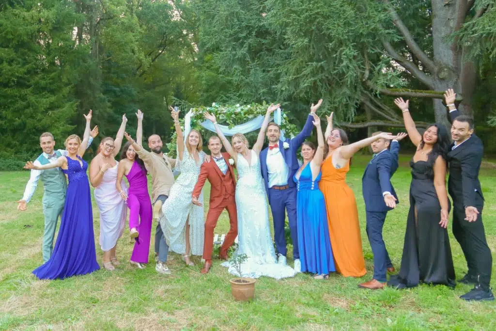 Mariés et invités posant bras levés devant une arche fleurie dans un parc